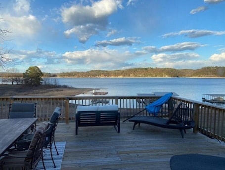 A wooden deck with a table and chairs, overlooking a lake with a dock and trees in the background.