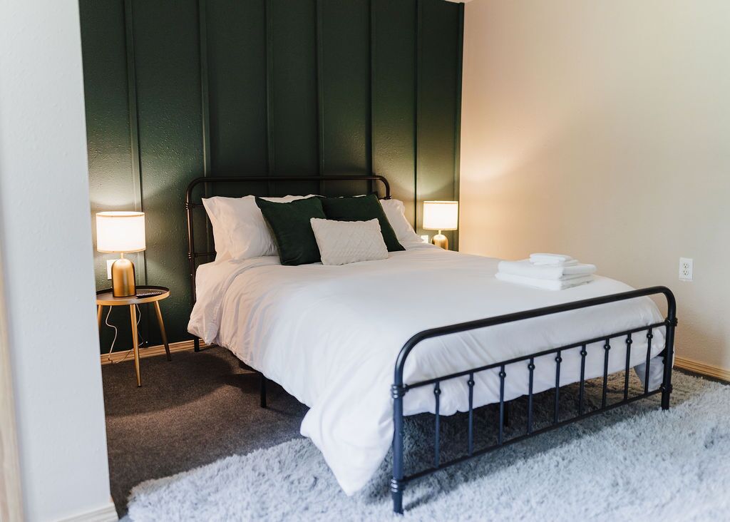 Una cama bien hecha con ropa de cama blanca y almohadas verdes en una habitación con paredes verde oscuro y cabecera de metal negro.