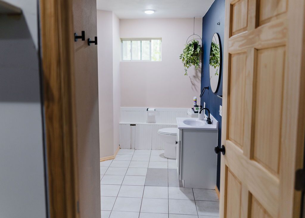 Une salle de bain moderne avec des toilettes, un lavabo et un miroir blancs. Une fenêtre laisse entrer la lumière naturelle et une plante en pot est posée sur le mur.