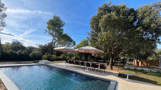A rectangular swimming pool with clear water, surrounded by a tiled deck. There are outdoor tables and chairs under umbrellas, and trees in the background. - Coeur de Lavande - Maison climatisée avec piscine (Fox-Amphoux)