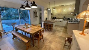 A modern kitchen with a central island, a dining area with wooden furniture, and a large window offering a view of the outdoors. - Coeur de Lavande - Maison climatisée avec piscine (Fox-Amphoux)