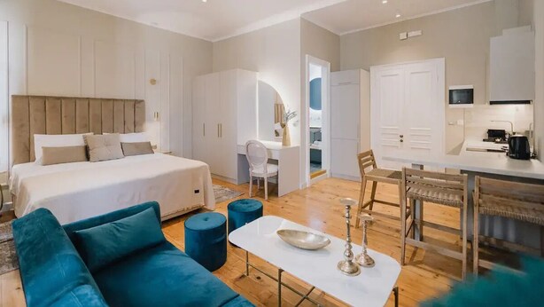 Ein modernes Hotelzimmer mit einem großen Bett, einer Sitzecke mit Sofa und Hocker, einem Esstisch mit Stühlen, einer Küchenzeile und einem durch einen Bogen sichtbaren Badezimmer.