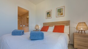 Una habitación de hotel con cama, dos almohadas, una mesita de noche con lámpara y un buró de madera.