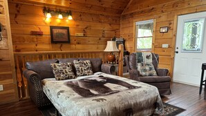 Interior de una cabaña de troncos con una cama grande, sillones de cuero y piso de madera.