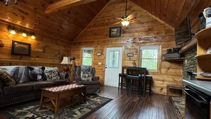 Interior acogedor de una cabaña con techo de madera, un sofá, una mesa de centro, una silla, una mesa de comedor y una zona de cocina.