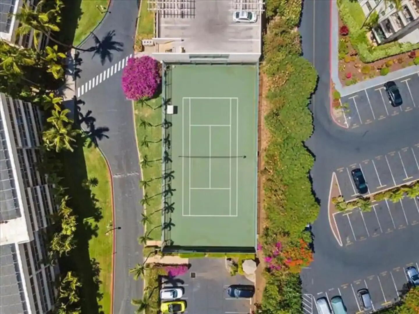 En vy över en tennisbana sedd från ovan, omgiven av parkeringsplatser och grönska.