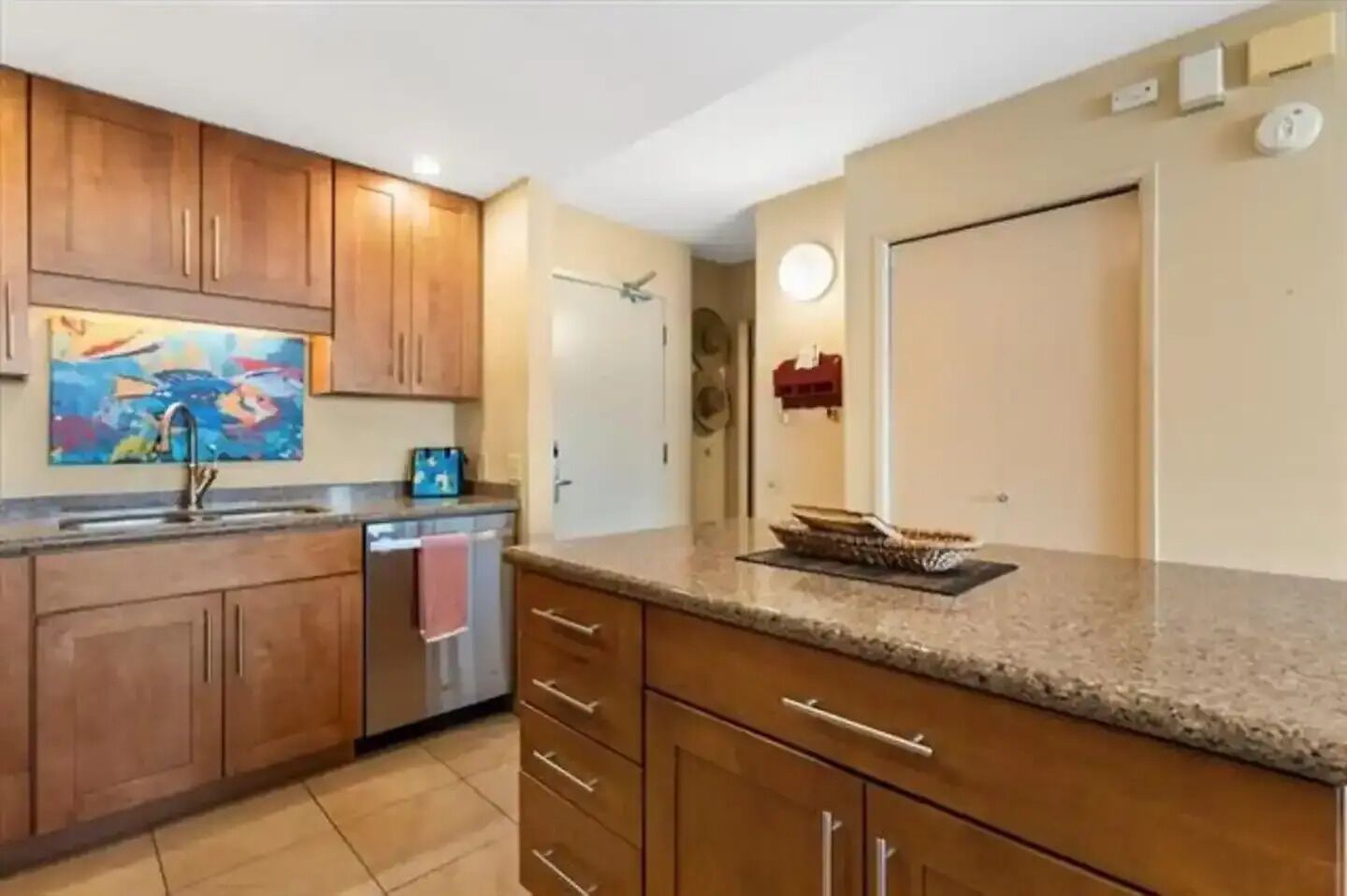 A kitchen with wooden cabinets, a stainless steel dishwasher, a granite countertop, and a painting on the wall.