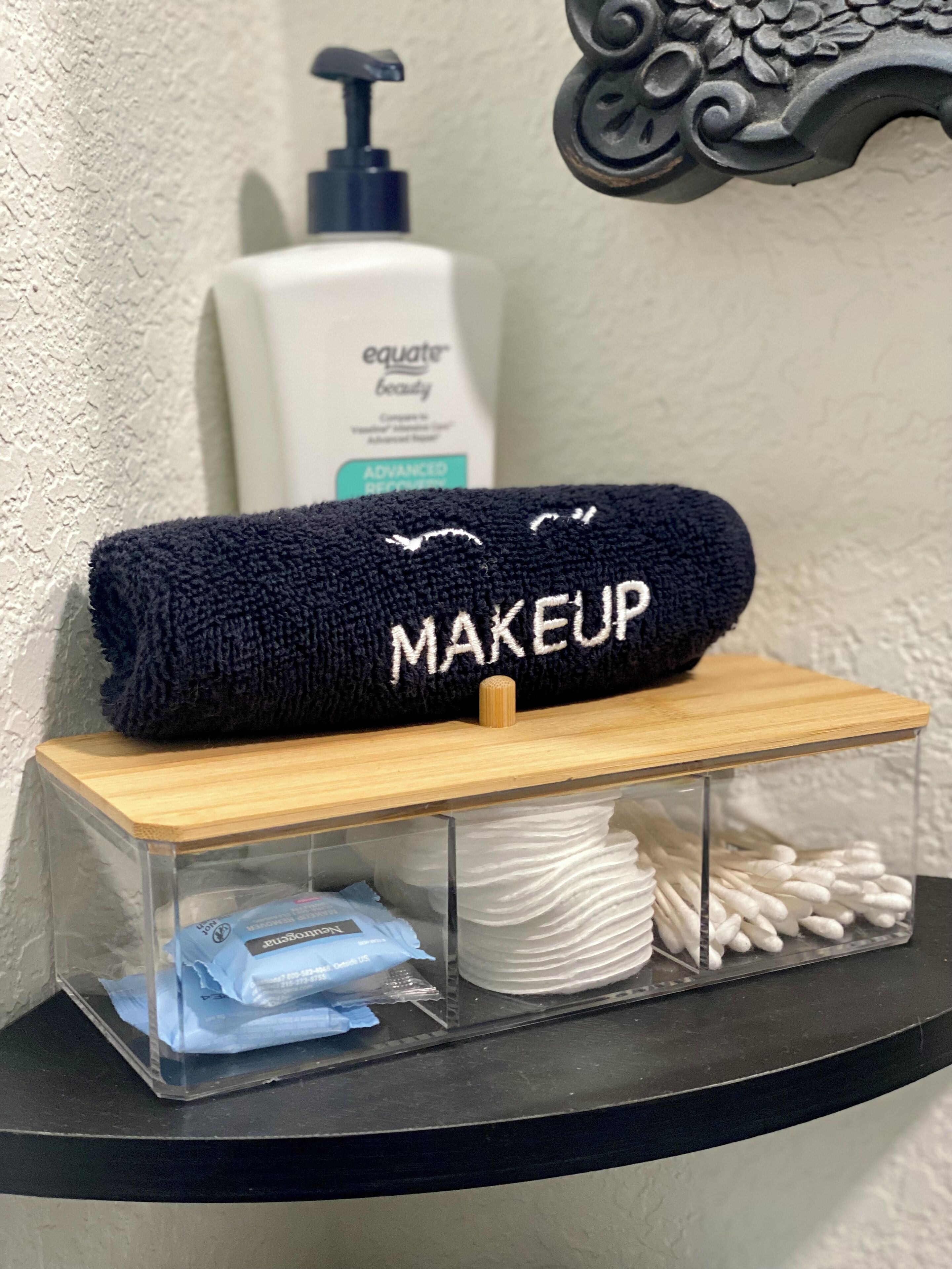 A bathroom vanity with a black towel labeled "MAKEUP" and a clear container holding cotton swabs and a blue tissue package.
