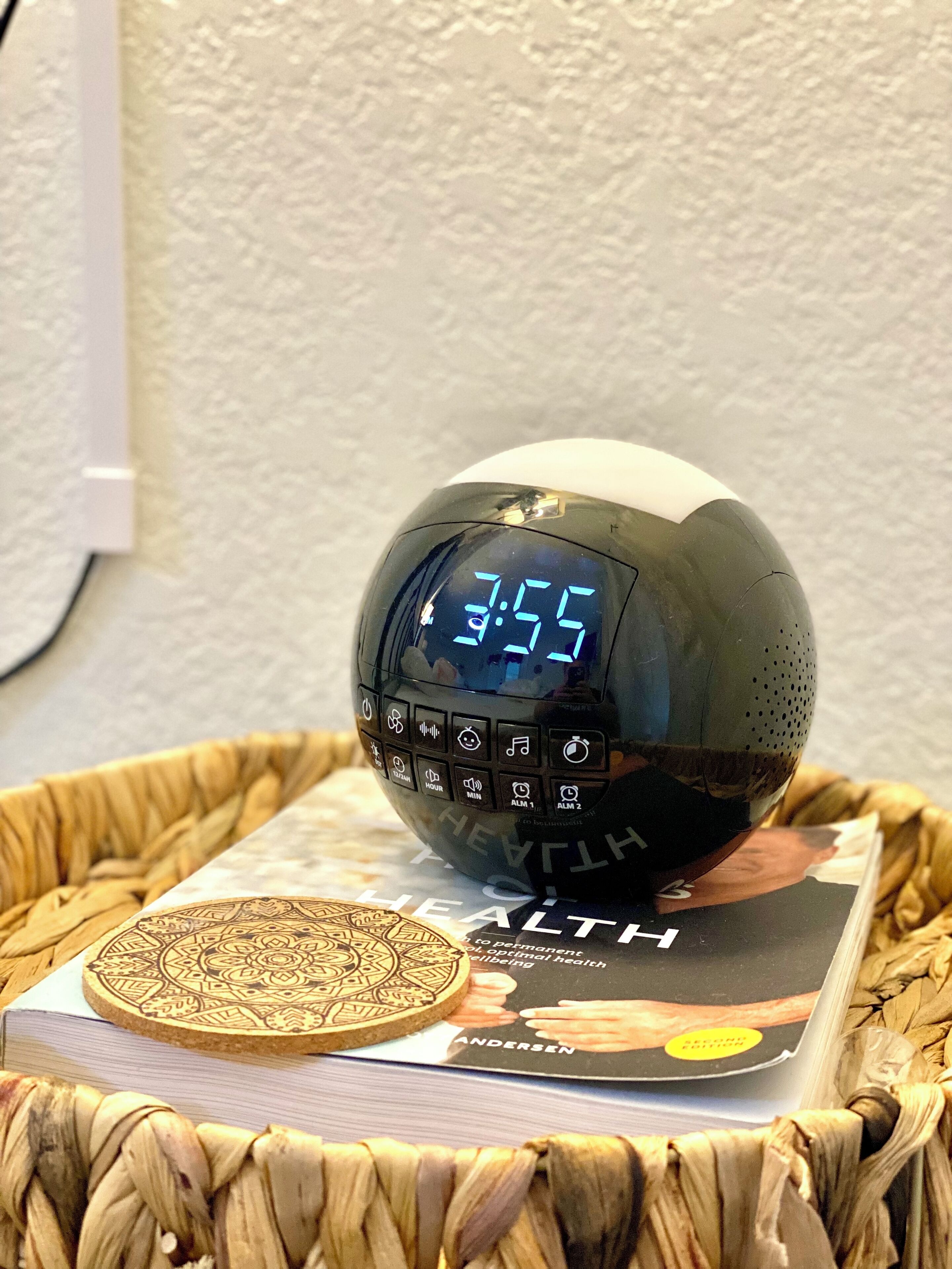Una sveglia digitale mostra l'ora 7:55, posizionata su un cesto di vimini contenente un libro intitolato “HEALTH”.