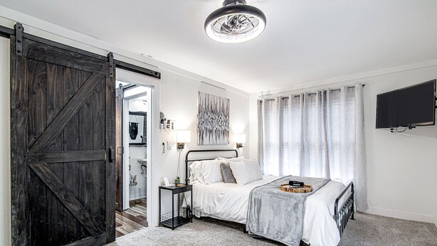 A bedroom with a bed, a nightstand, a large wooden door, a ceiling light, and a TV mounted on the wall.