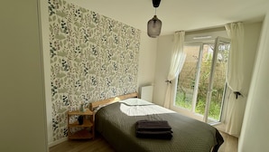 A bedroom with a large window, a bed with a green bedspread, a bedside table with a lamp, and a wall with a leafy pattern.
