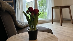 A living room with a round wooden table, a sofa, a potted plant with red flowers, and a window with blinds.