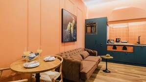 A cozy room with a brown sofa, a round wooden table with a glass of orange juice, and a kitchen area with orange tiles and a blue cabinet. - Mantzarou #6 - Corfu Town Luxury Suites

 (Kerkira)