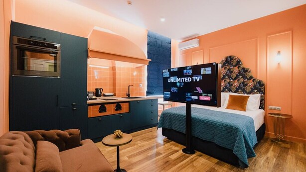 A modern hotel room with a kitchenette, a bed with a blue bedspread, a TV displaying 'UNLIMITED TV', and a small round table. - Mantzarou #6 - Corfu Town Luxury Suites

 (Kerkira)