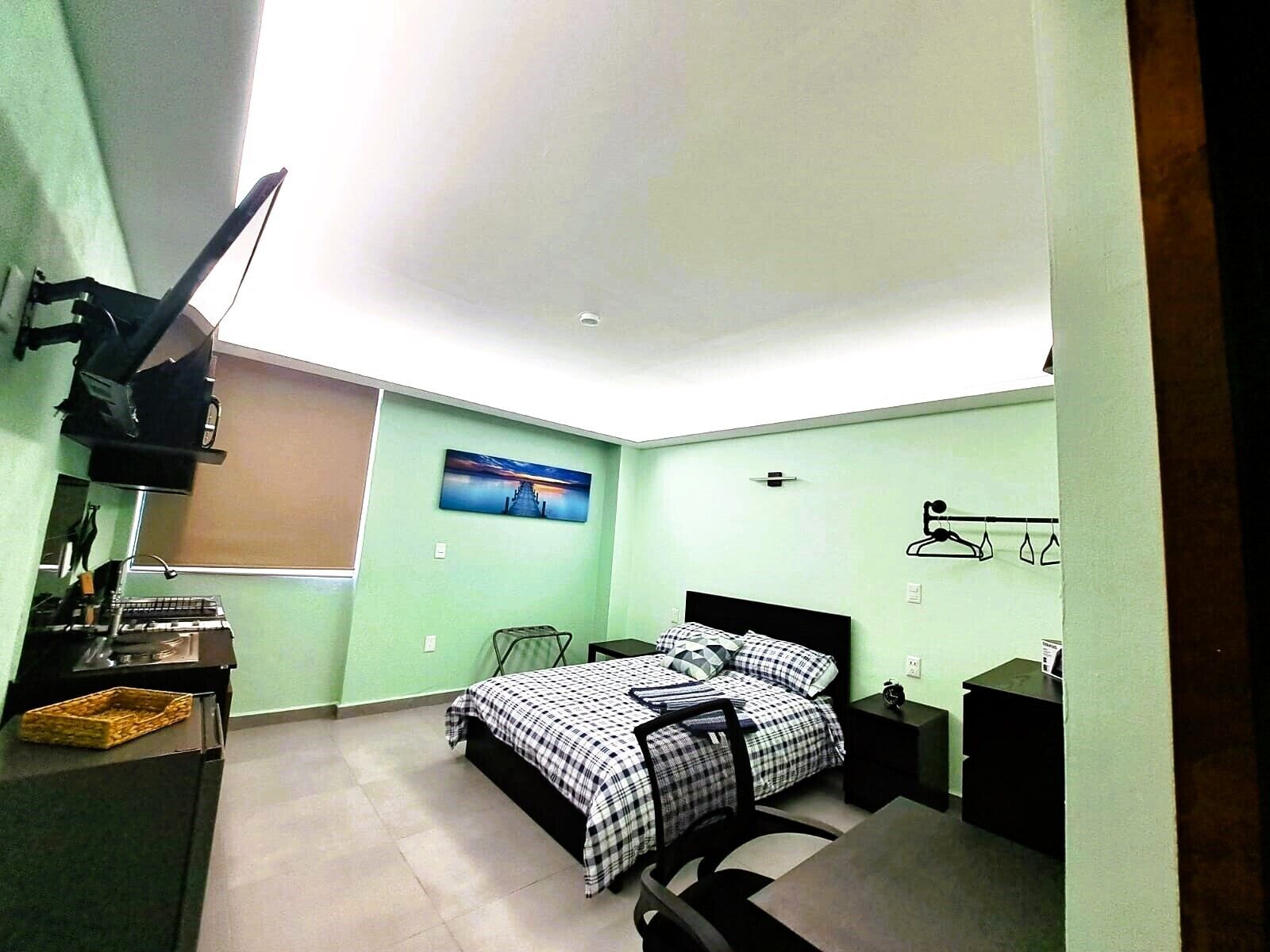 A small, well-lit bedroom with a bed, a desk, a chair, a hanging light fixture, and a wall-mounted shelf.
