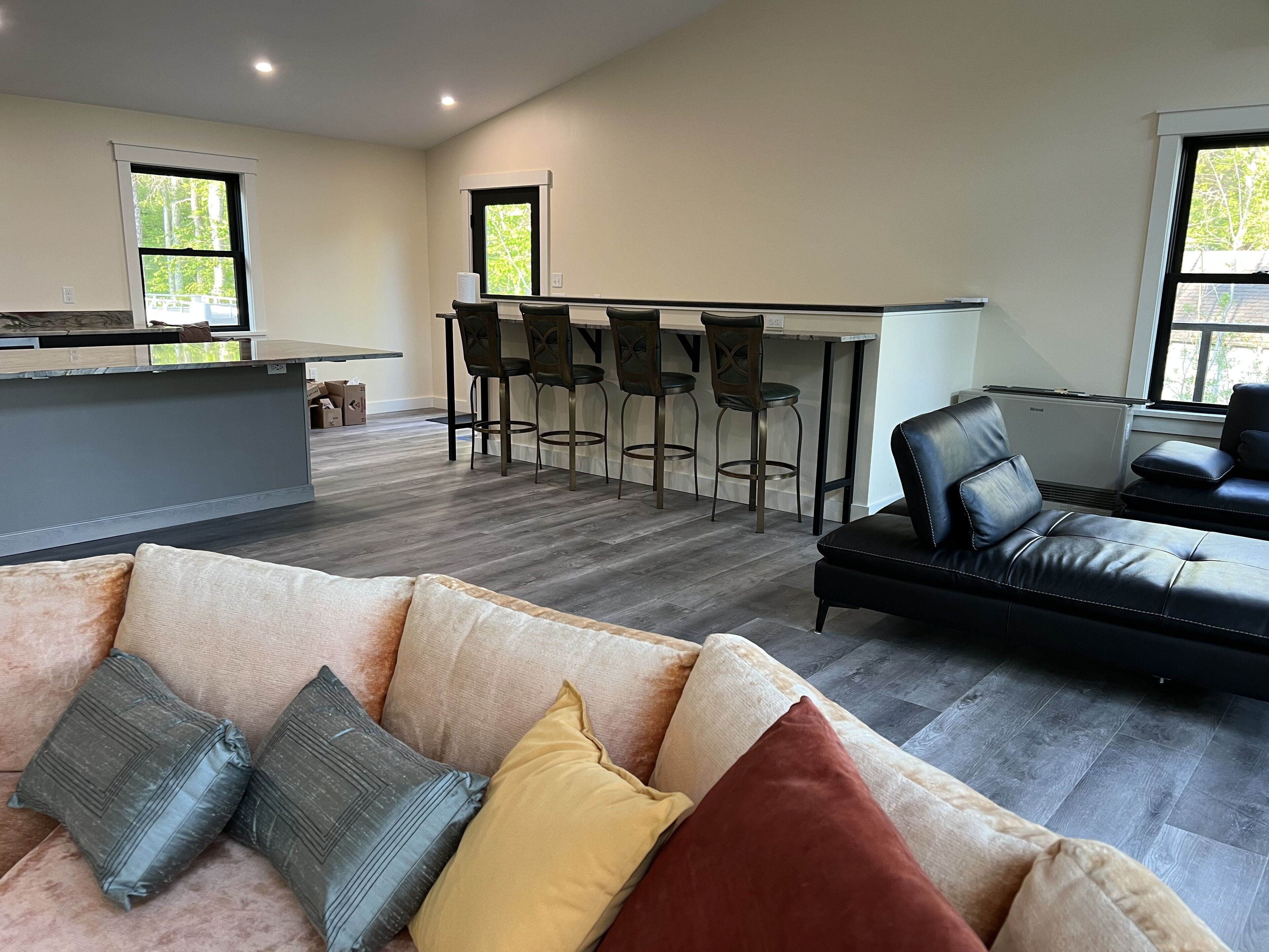 Ein modernes Wohnzimmer mit einem grauen Sofa, einem schwarzen Ledersofa und einem beigefarbenen Schlafsofa mit Kissen. Es gibt eine Küchenzeile mit einer Theke und Barhocker.