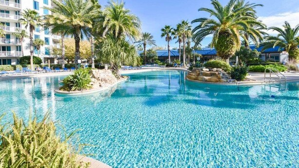 Une grande piscine extérieure à l’eau d’un bleu cristallin, entourée de palmiers et bordée d’un immeuble à plusieurs étages doté de balcons.