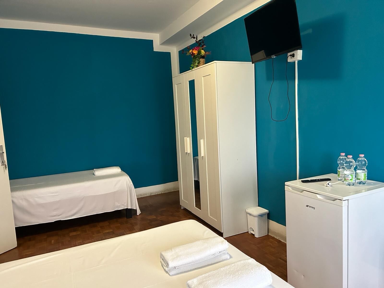 A room with teal walls, a bed with white linens, a wardrobe, a small fridge, and a TV mounted on the wall.