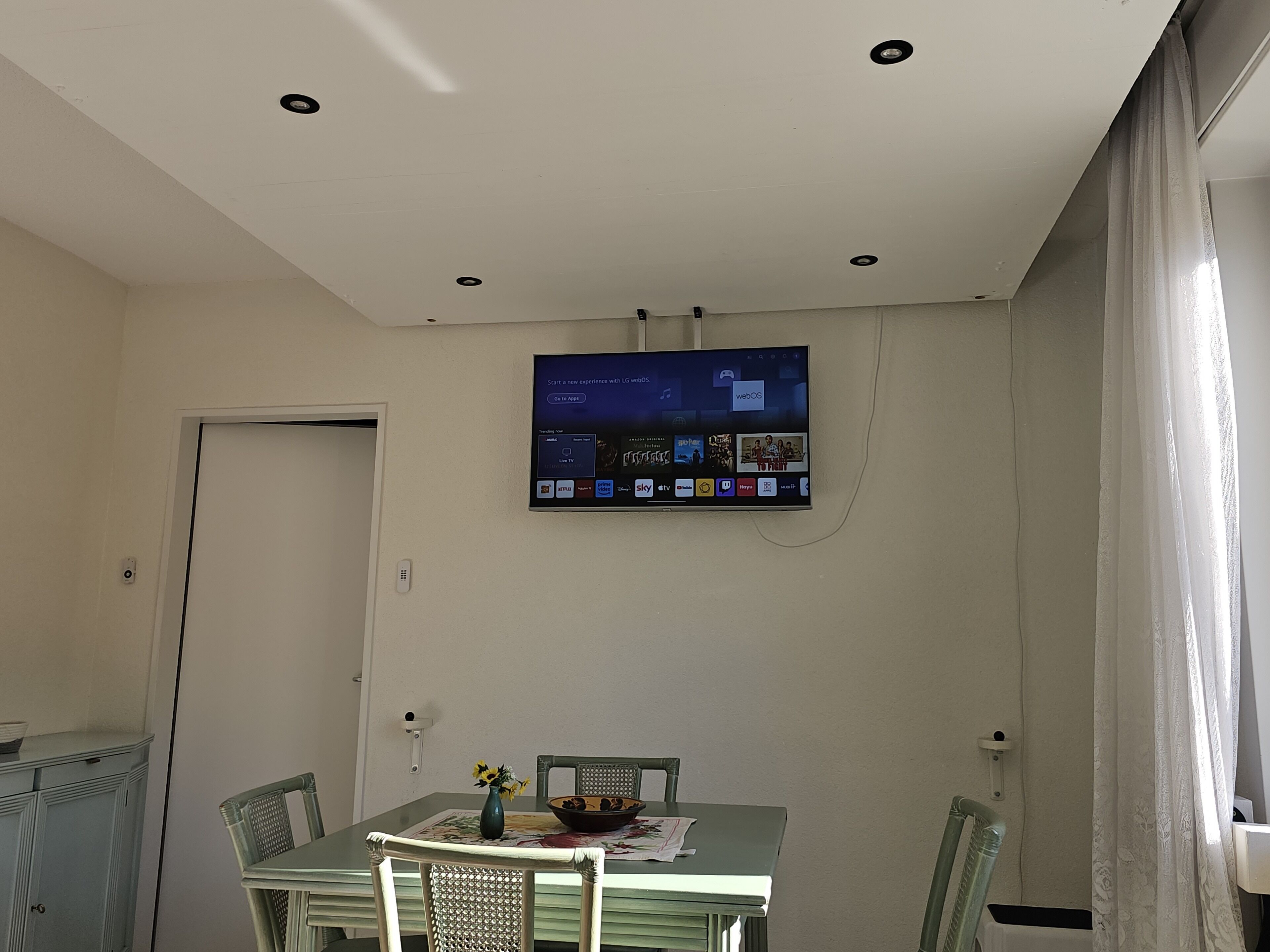 Espaço de refeições com mesa, cadeiras, um vaso de flores e uma televisão fixada na parede exibindo vários aplicativos.