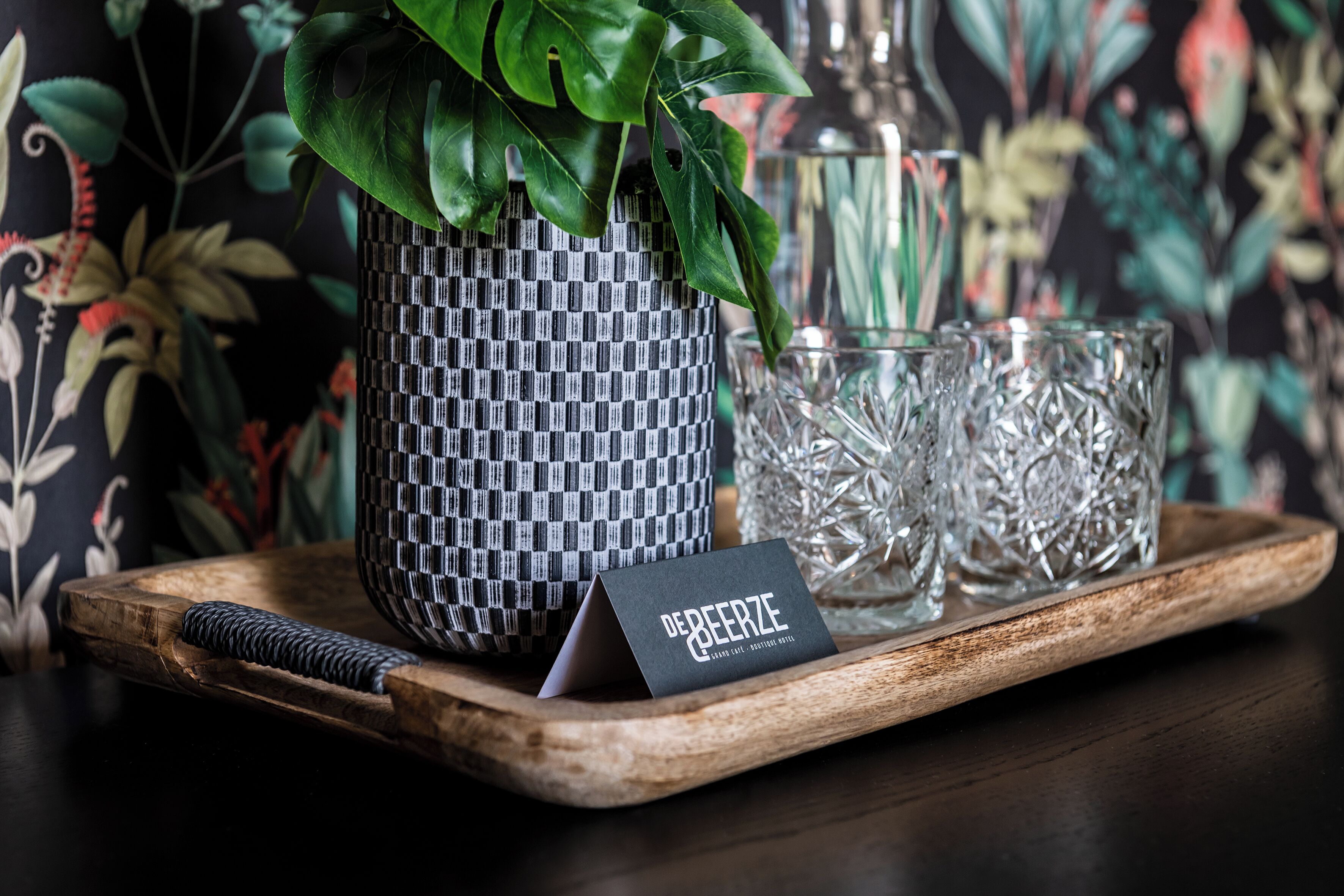 Un plateau avec un pot de fleurs en fibres tressées, deux verres en cristal et une carte noire avec du texte.