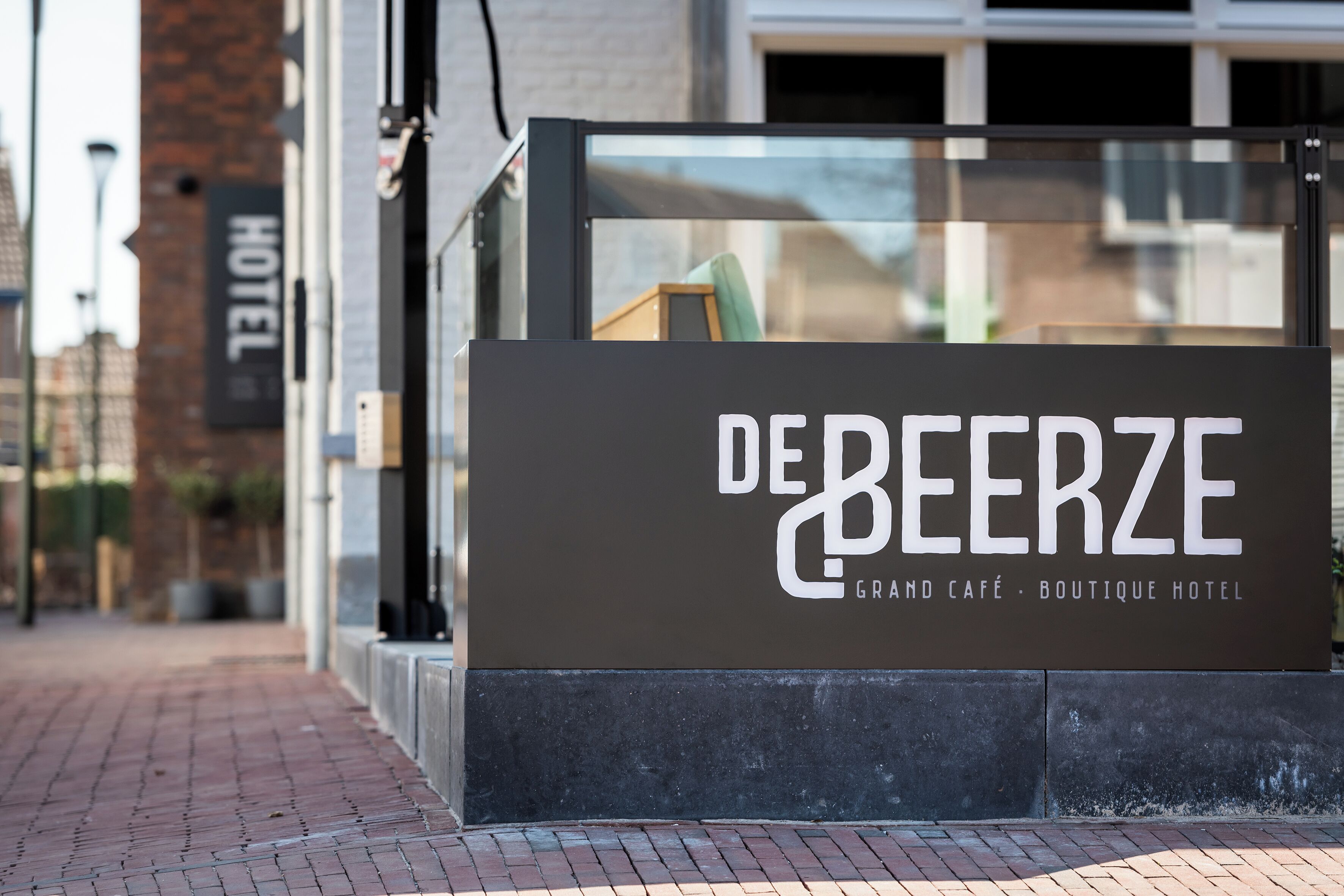 A sign for De Beerze Grand Café - Boutique Hotel with a clear view of the building's entrance and a brick wall in the background.