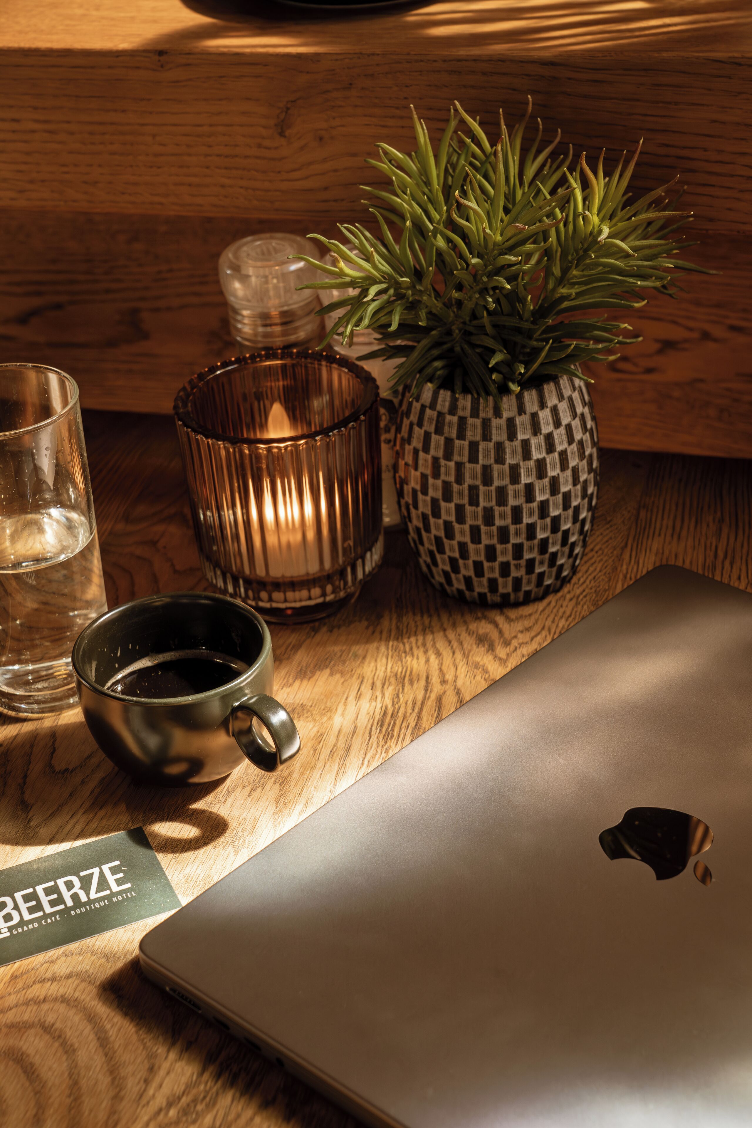 Une table en bois sur laquelle se trouvent un ordinateur portable, un verre d’eau, une tasse, une bougie et un pot à carreaux.