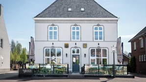Een wit gebouw met een grijs dak, een blauwe deur en twee kleine ramen boven de deur. Twee parasols op stokken staan voor het gebouw.