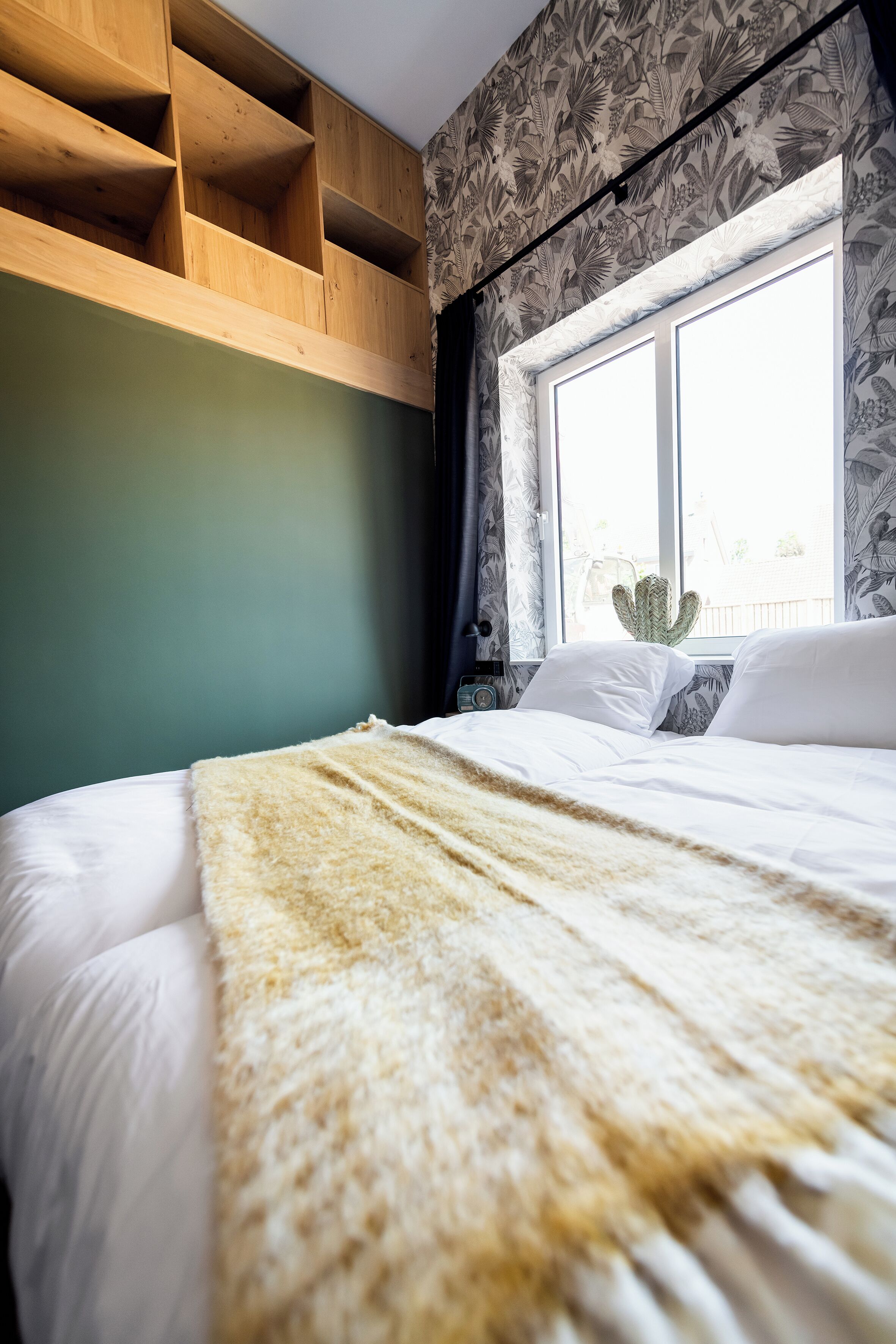 Ein Schlafzimmer mit einem Bett, einem Fenster mit Vorhängen, einer Wand mit Blumenmuster und einem Holzbücherregal.