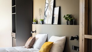 Una cama bien hecha con almohadas blancas y mostaza, un estante en la pared con plantas y obras de arte enmarcadas, y un marco de puerta de madera.