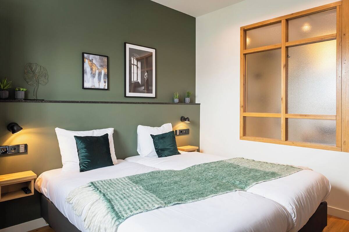 Un dormitorio moderno con una cama grande, dos mesitas de noche, una mesita auxiliar, una pared verde y un ventanal con vidrio esmerilado.