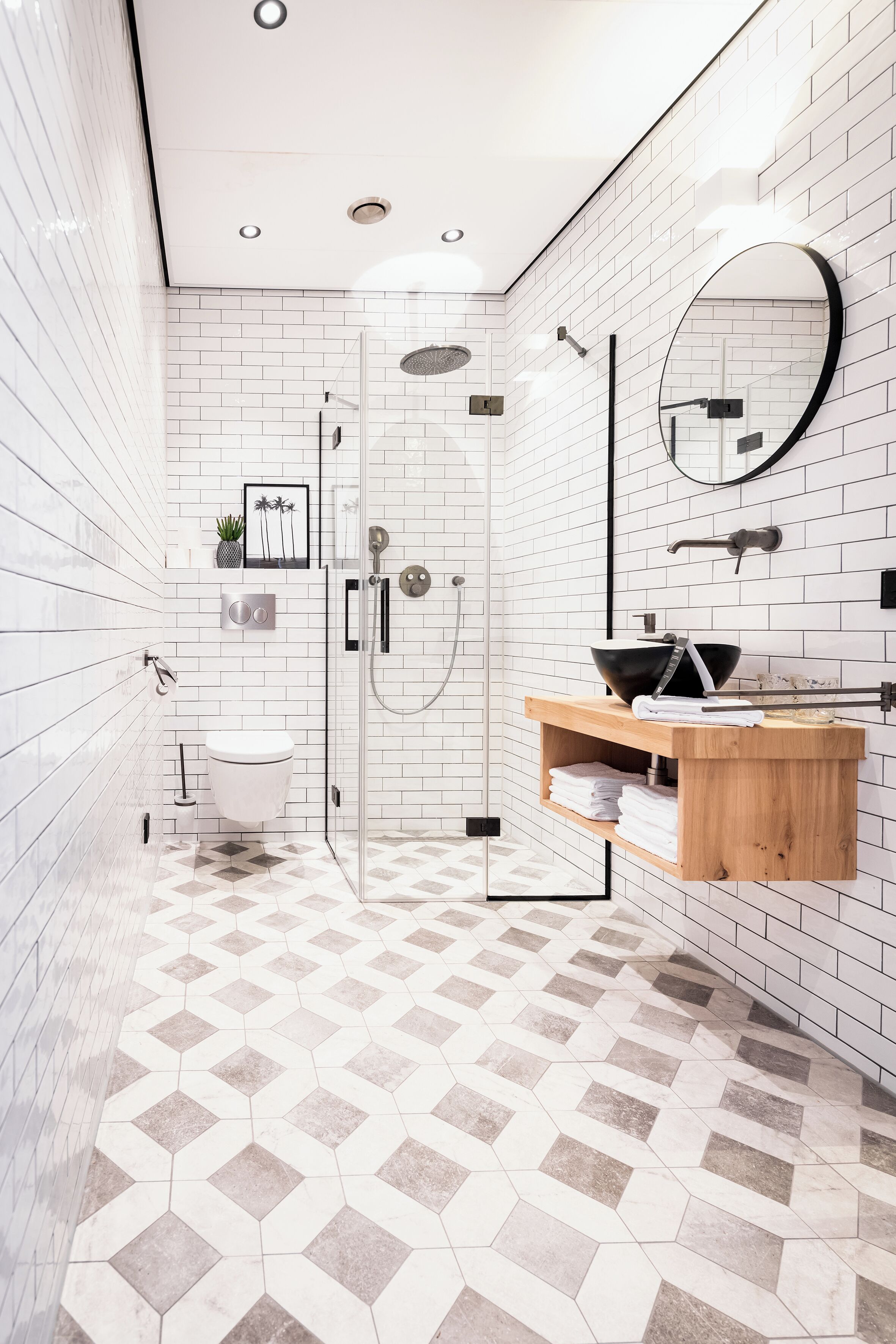 Un baño moderno con ducha envuelta en vidrio, tocador de madera con lavabo negro y suelo de baldosas blancas con diseño geométrico.