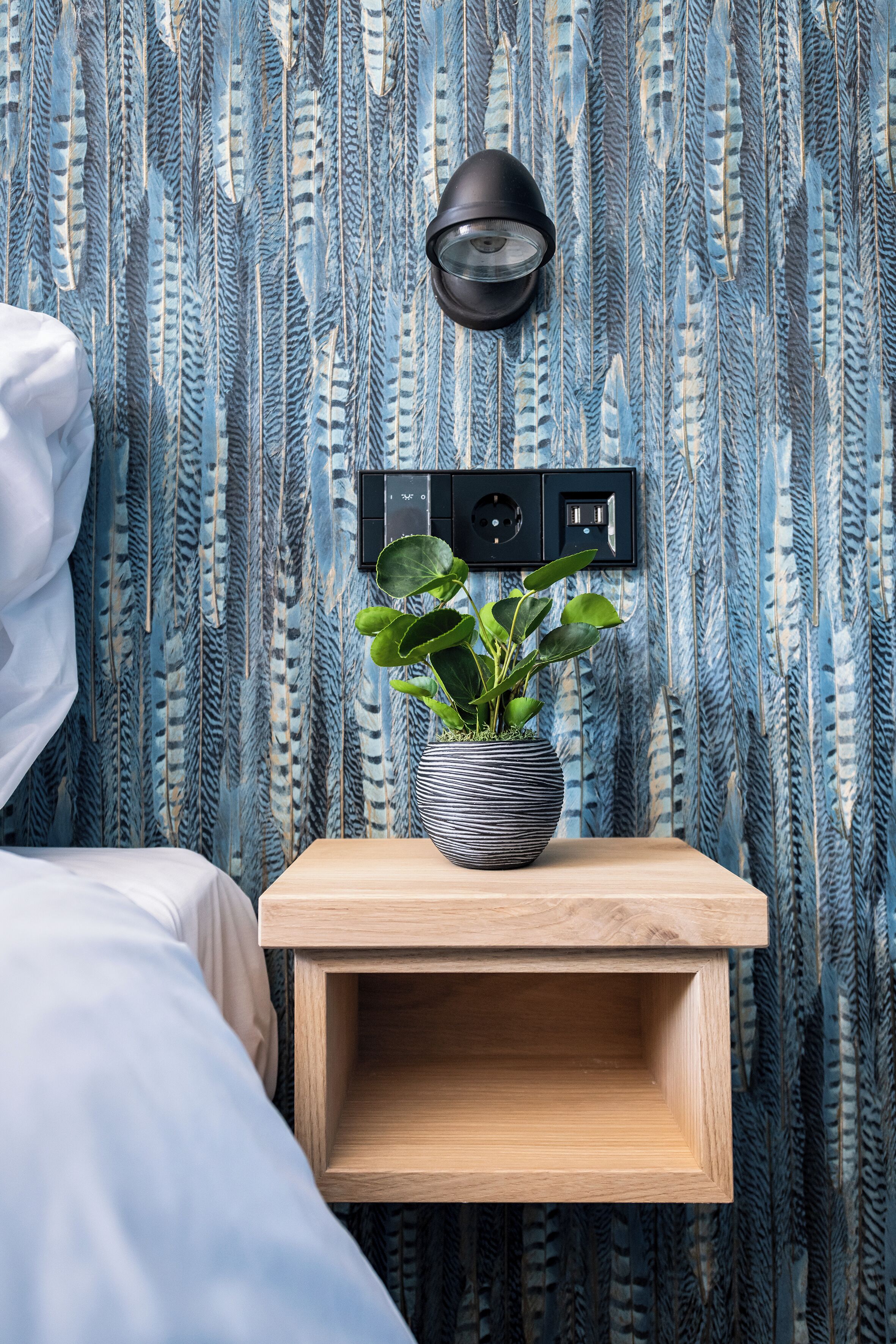 Una cama bien hecha con ropa de cama blanca, una mesita de noche con una planta en maceta y una lámpara de pared.