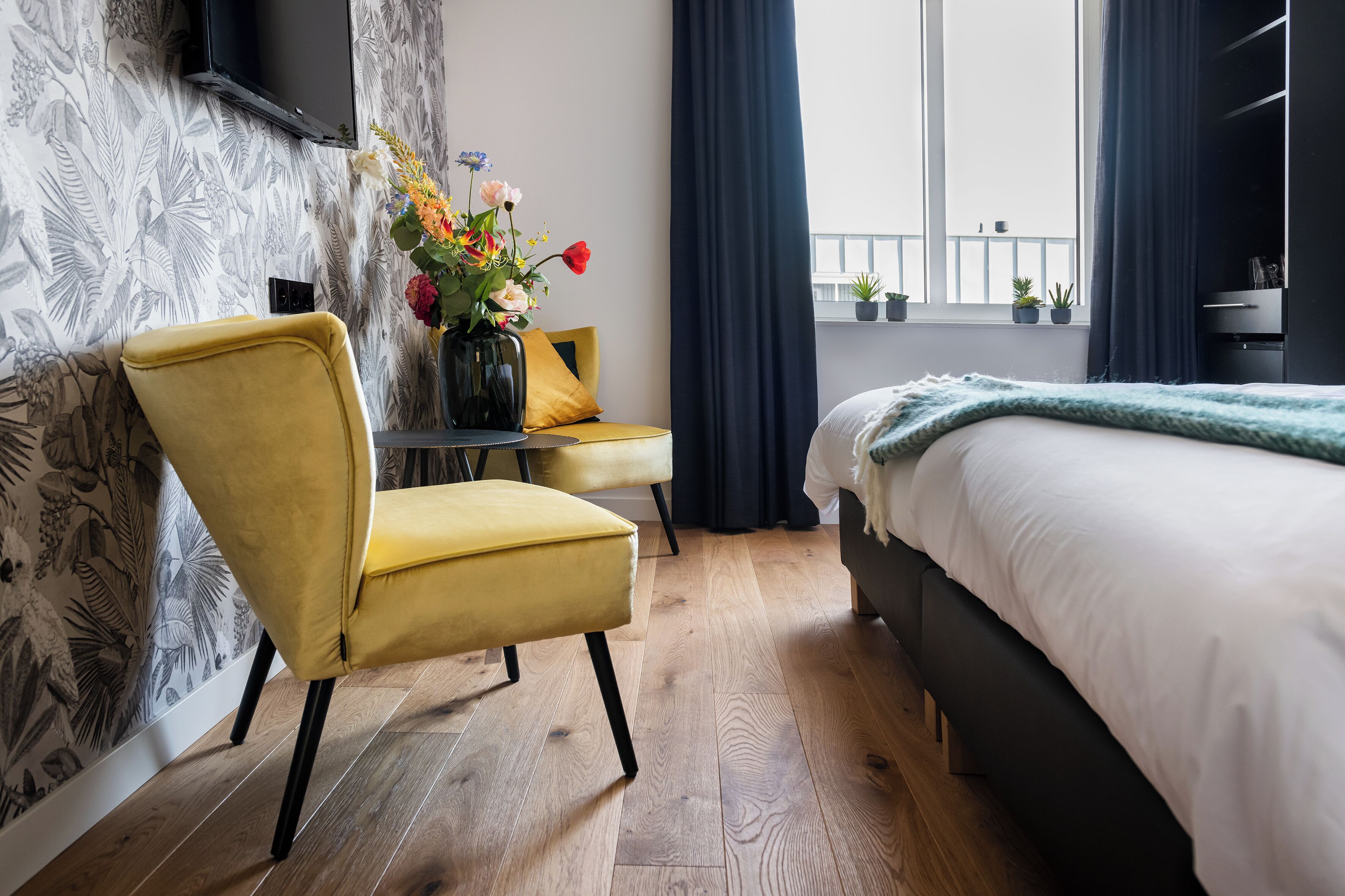Un dormitorio moderno con suelo de madera, una cama con ropa de cama blanca, un sillón amarillo, una mesita con un florero y una ventana con cortinas.