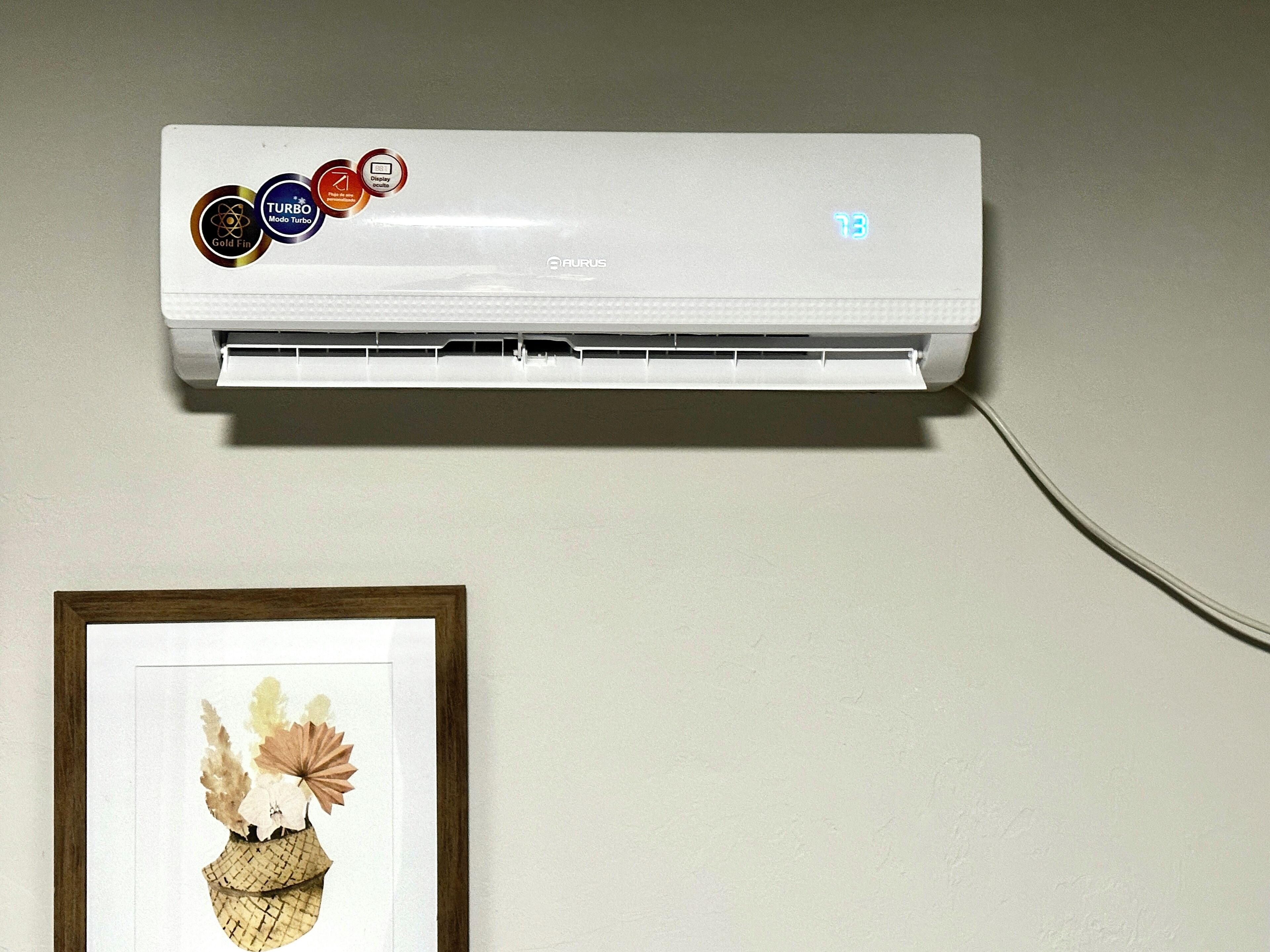 Un climatiseur mural doté d’un écran numérique, d’un câble d’alimentation et d’un cadre contenant une image de fleurs placé en dessous.
