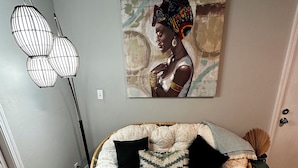 Una habitación con una zona de estar acolchada, una lámpara de pie, un cuadro, una mesita con un mando a distancia y un jarrón decorativo.