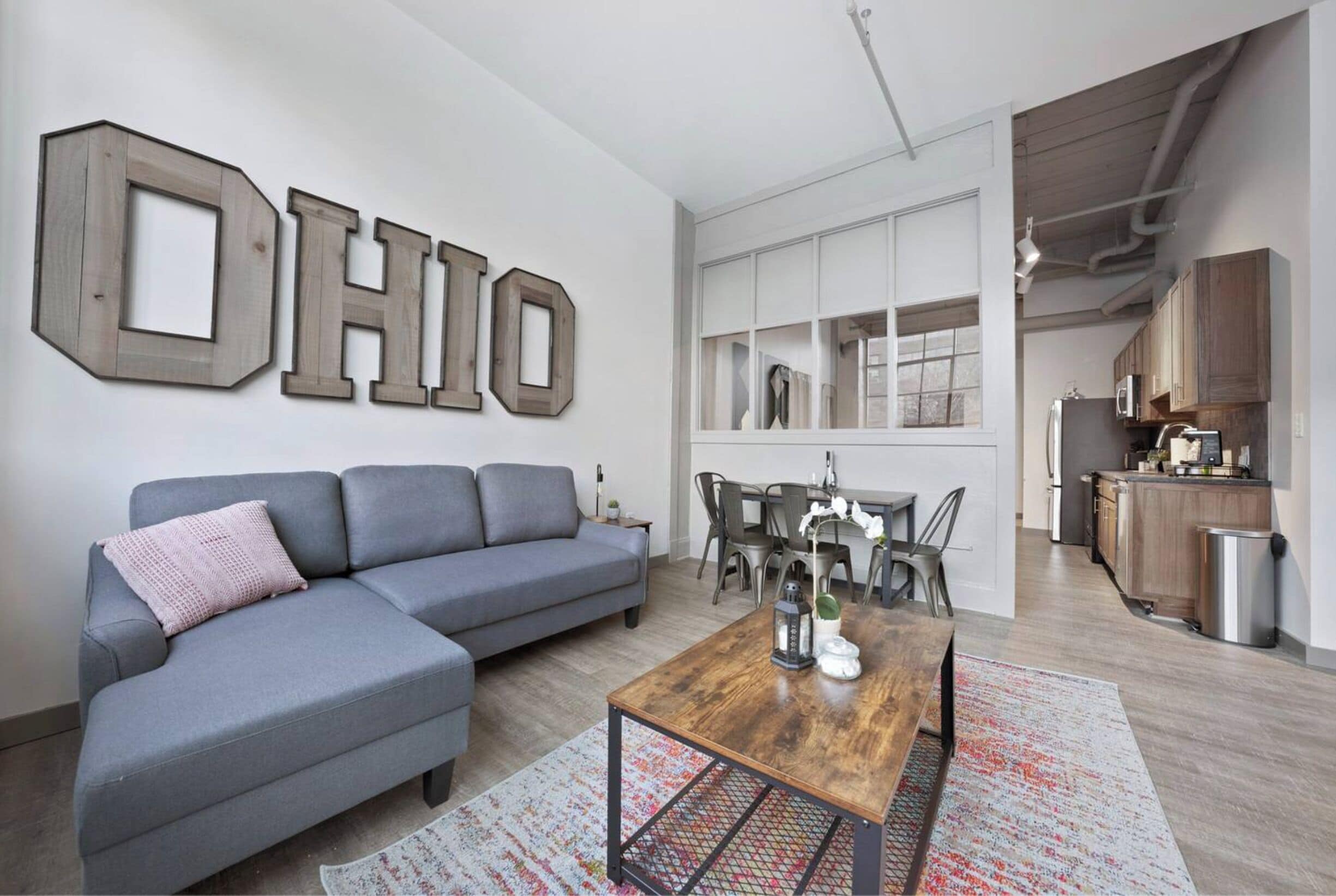 A modern living room with a gray sectional sofa, a wooden coffee table, and a dining area with a white table and chairs. A large "OHIO" sign decorates the wall.