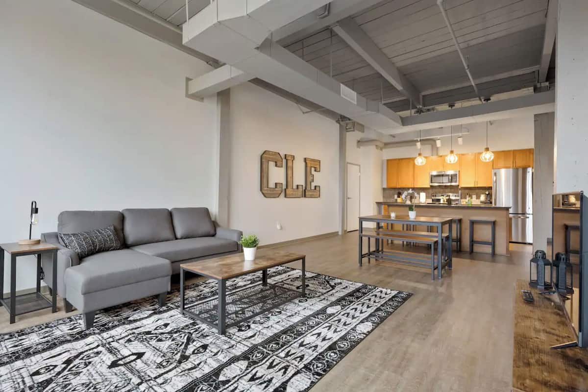 A modern living room with a gray sofa, coffee table, and patterned rug. A kitchen with wooden cabinets and stainless steel appliances is visible in the background.