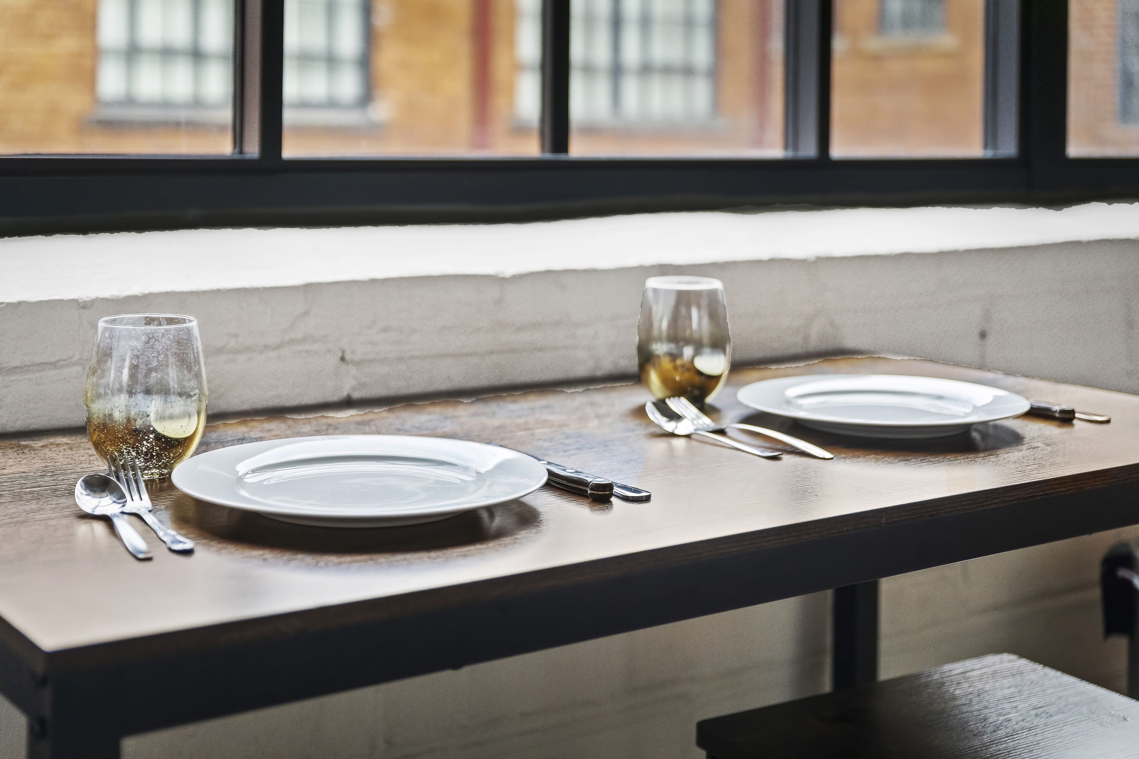 Une table à manger, dressée avec des assiettes, des couverts et des verres, placée devant une fenêtre offrant une vue sur des immeubles.