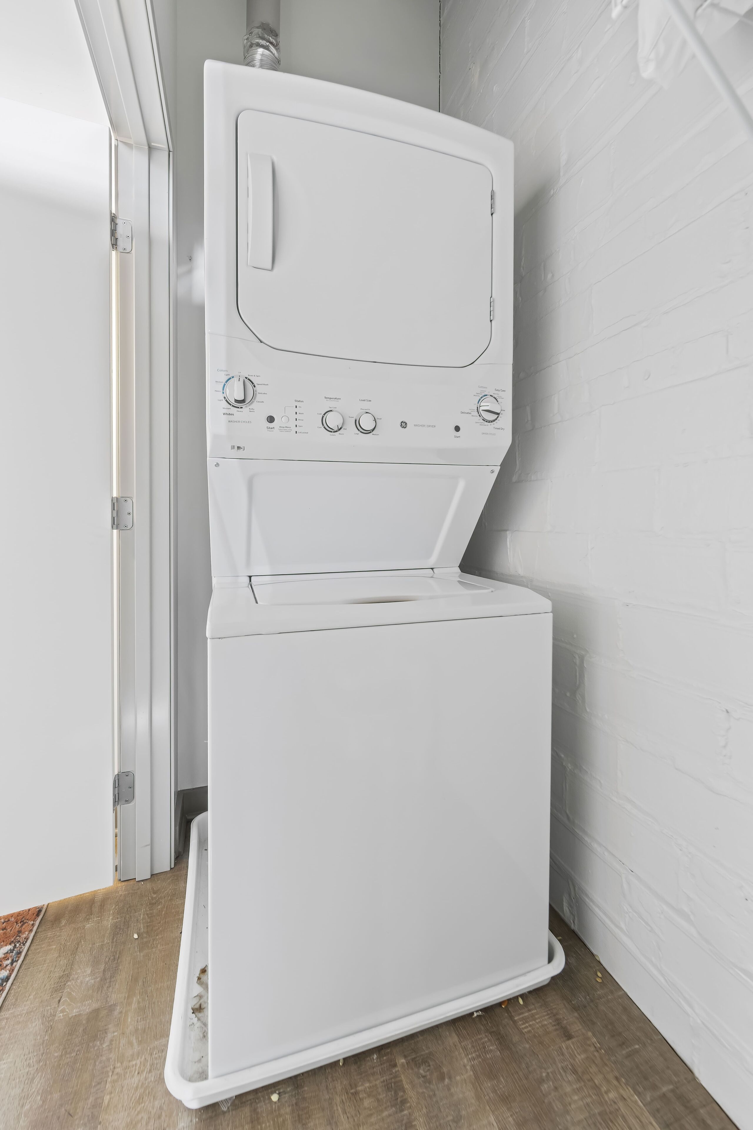 Een witte wasmachine in een hoek van een kamer met een houten vloer en witte bakstenen muren.