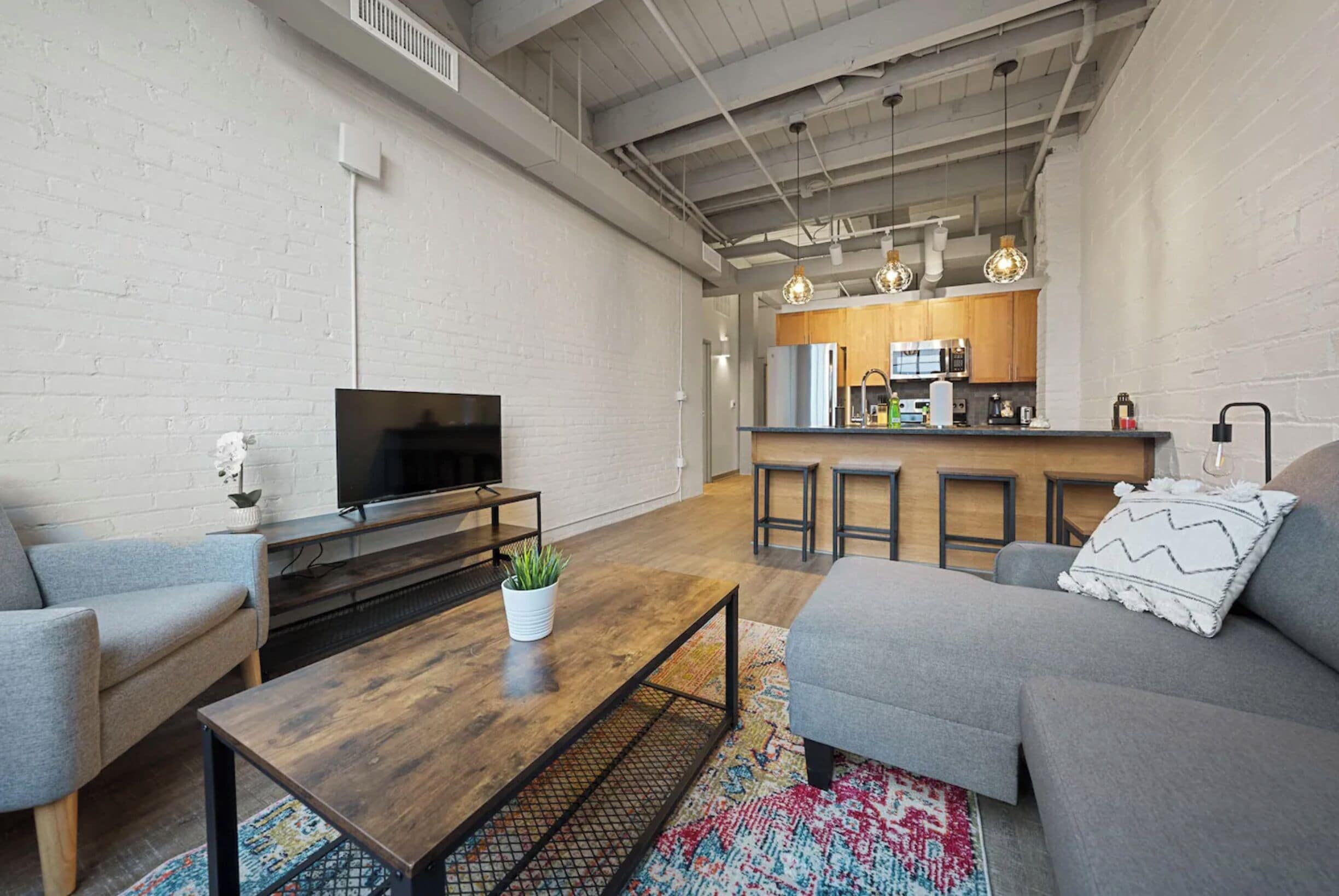 A modern living room with a flat-screen TV, a coffee table with a plant, and a gray sofa. A kitchen with wooden cabinets and a bar area is visible in the background.