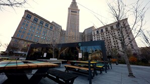 An outdoor seating area with tables and benches, a modern building with a glass facade, and a tall building in the background. - 2125 Superior Living (Cleveland)