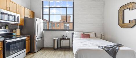 Una cocina compacta con armarios de madera, un refrigerador de acero inoxidable y un microondas. Hay un ventanal con vistas a edificios.