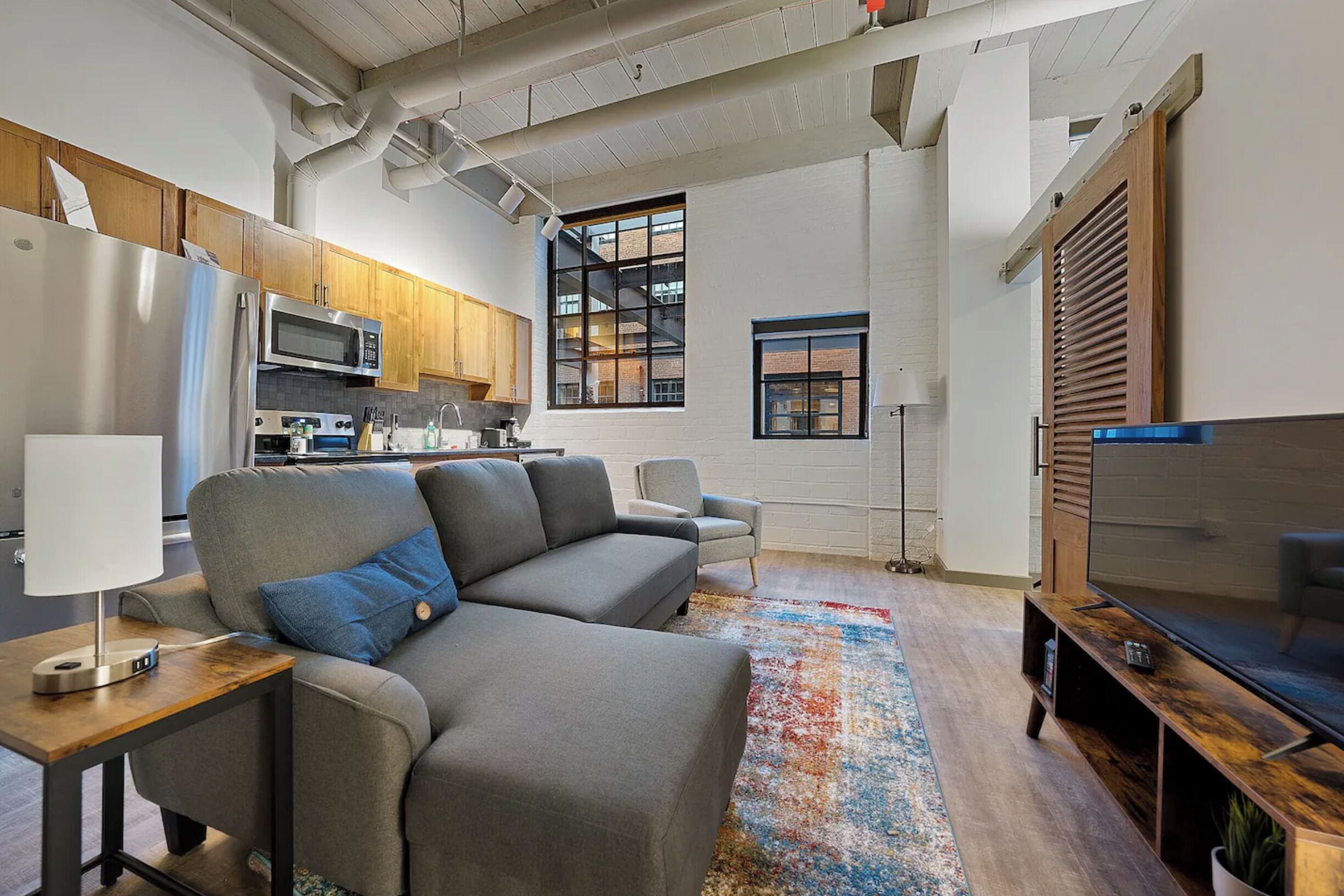 Una sala de estar moderna con un sofá gris, un mueble para TV y una cocina con armarios de madera y un microondas.