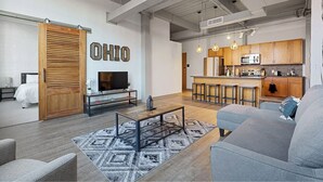 A modern living room with a grey sofa, a coffee table, and a TV. There is a kitchen area with wooden cabinets and a bar. - 2125 Superior Living (Cleveland)