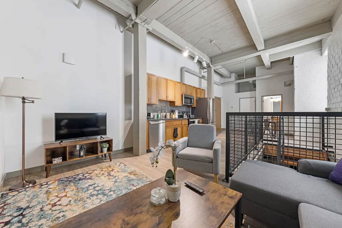 A modern living room with a TV, sofa, and coffee table, a kitchen area with cabinets and appliances, and a staircase leading to an upper level.