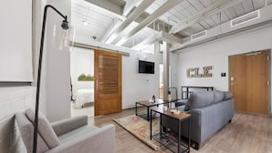 A modern living room with a sofa, a coffee table, and a lamp. There is a television on the wall and a door labeled "CLE". - 2125 Superior Living (Cleveland)