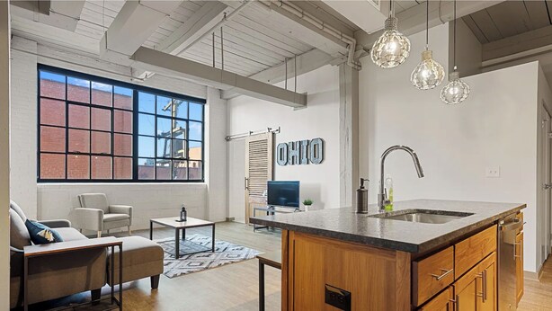 A modern kitchen with a large island, wooden cabinets, and a farmhouse sink. There is a dining area with a sofa and chairs, and a large window with a view of the outside. - 2125 Superior Living (Cleveland)
