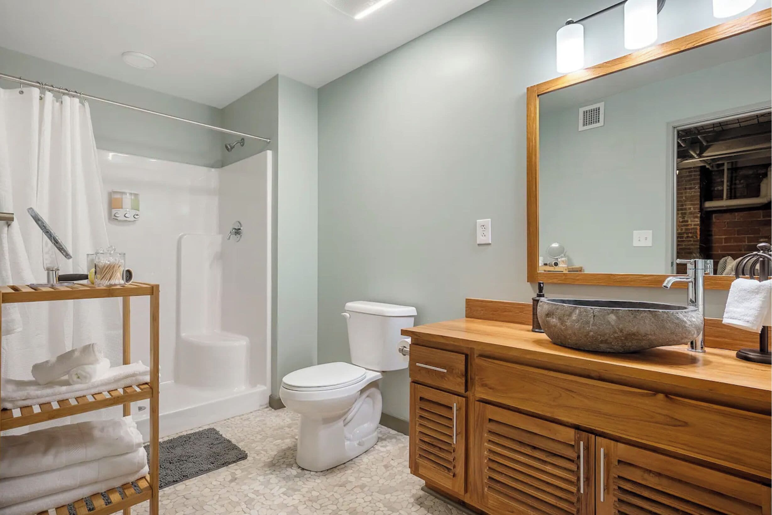 A modern bathroom with a walk-in shower, a toilet, a wooden vanity with a large mirror, and a shelf with towels.