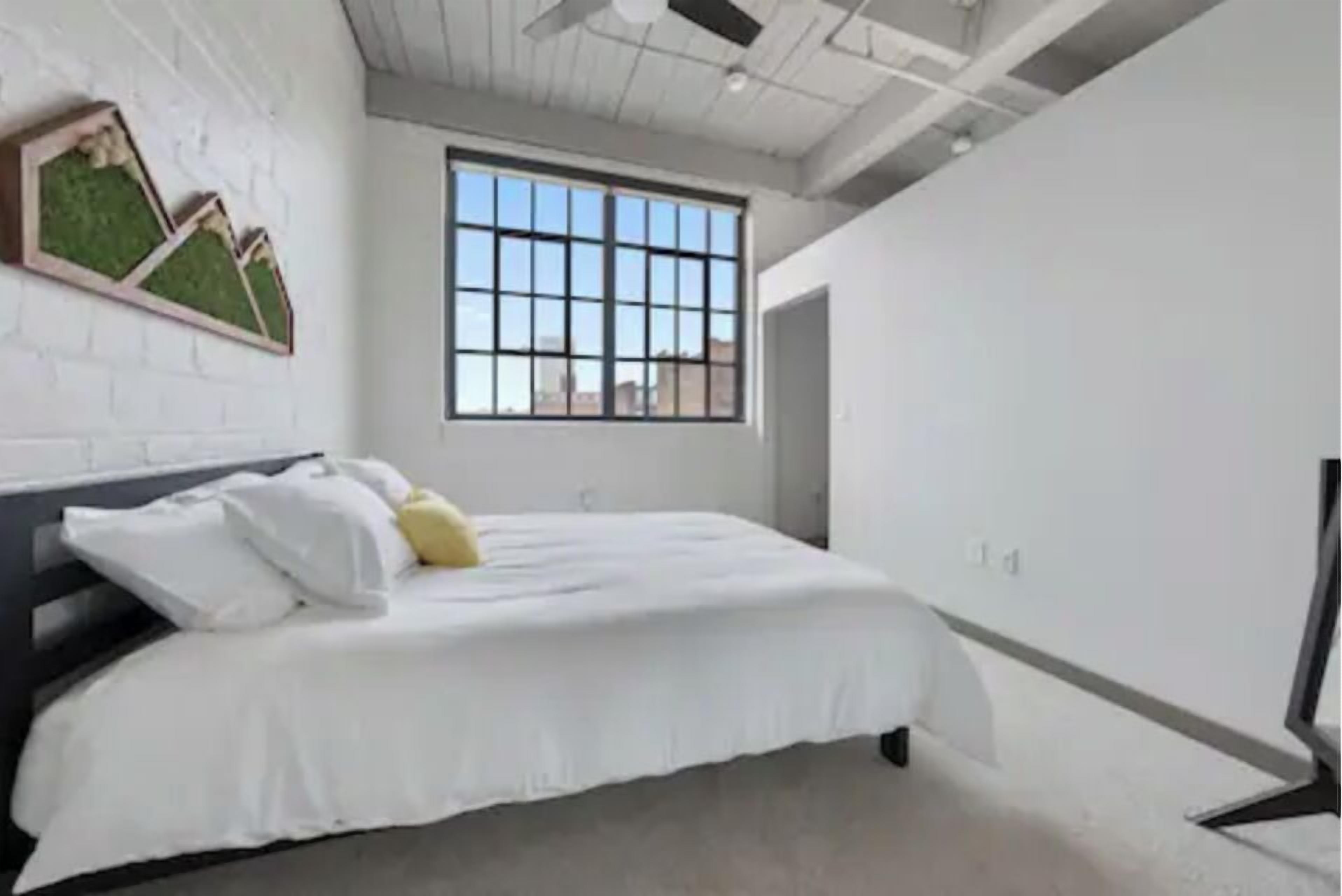 A modern bedroom with a large window, a bed with white bedding, and a wall-mounted artwork.