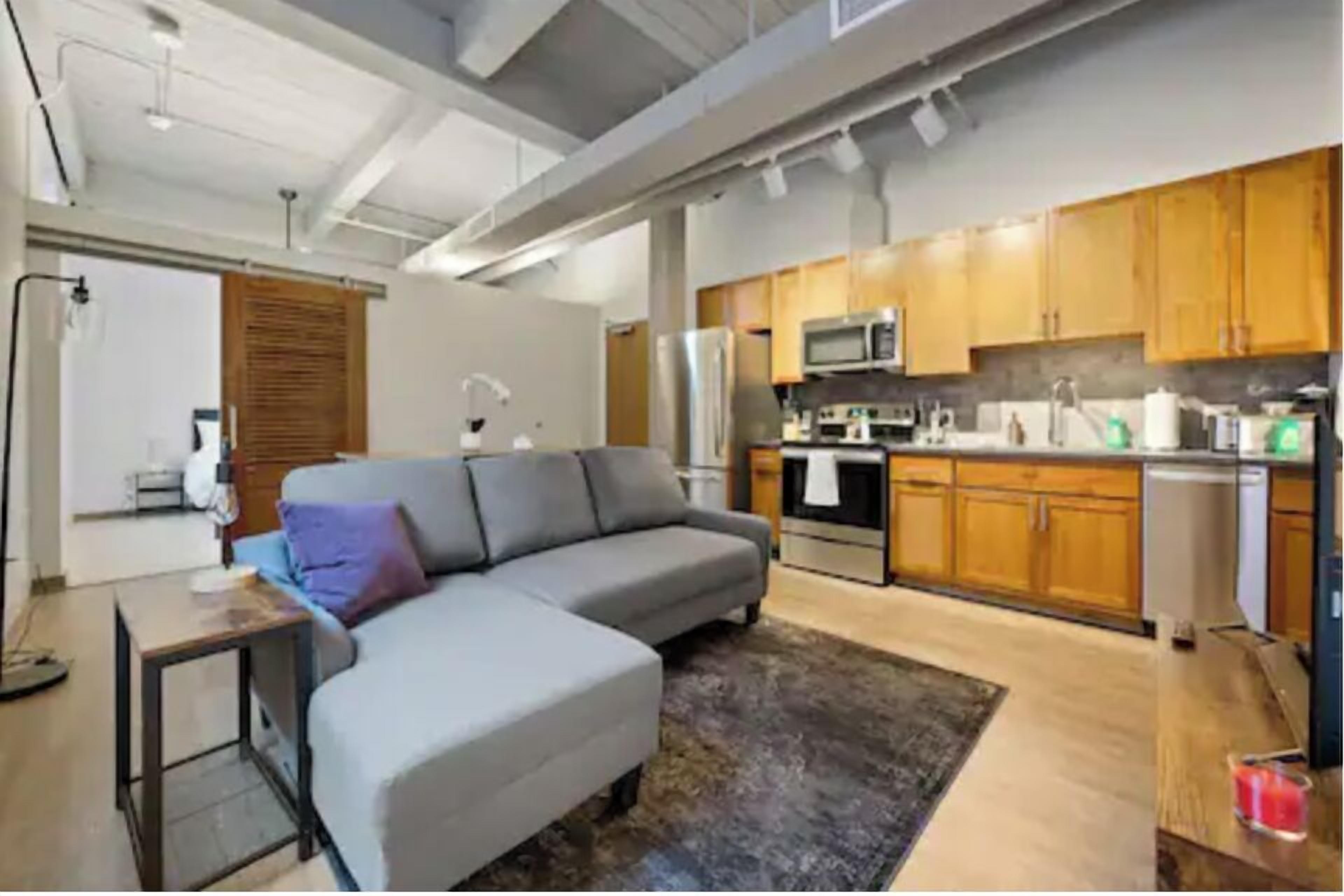 A modern kitchen with wooden cabinets, stainless steel appliances, and a grey sectional sofa in a living area.